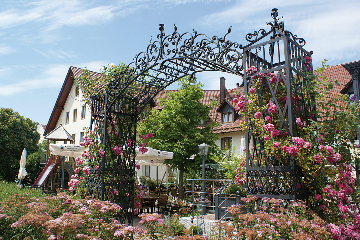 Ein großer schmiedeeiserner Torbogen mit Rosen und dem Gastgarten im Hintergrund