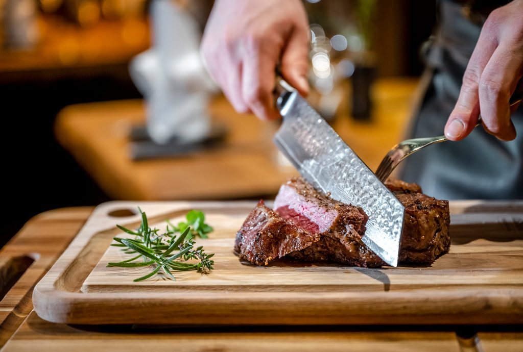 Raspls Genuss Schmiede Holzbrett mit Fleischstück wird von Messer in Scheiben geschnitten