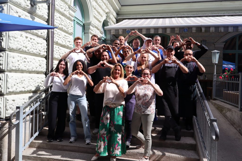 Mitarbeiter des Hofbräukeller auf einem Gruppenbild vorm Gebäude zeigen Herzen mit den Händen